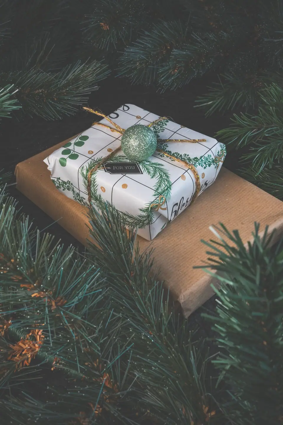 A small gift in printed paper and twine with a glitter ornament, resting on a kraft-paper parcel among long-needled pine branches